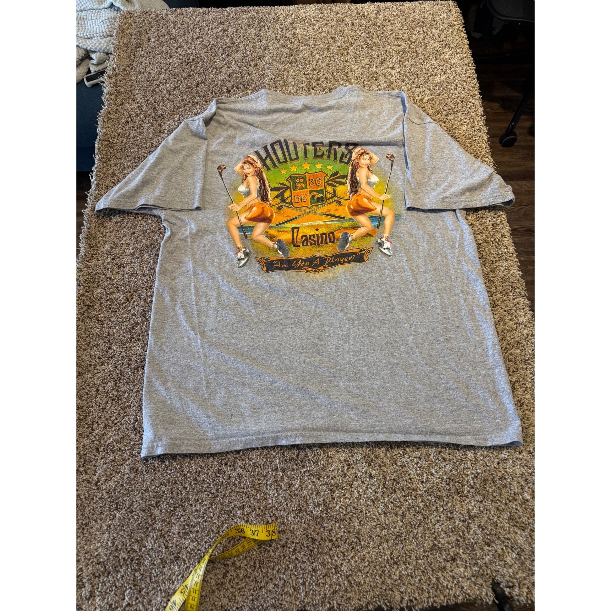 A heather gray short-sleeve t-shirt with a colorful graphic print of two pin-up girls and the words 'HOUTERS' and 'Las Vegas', laid flat on a brown carpet with a yellow measuring tape visible at the bottom.