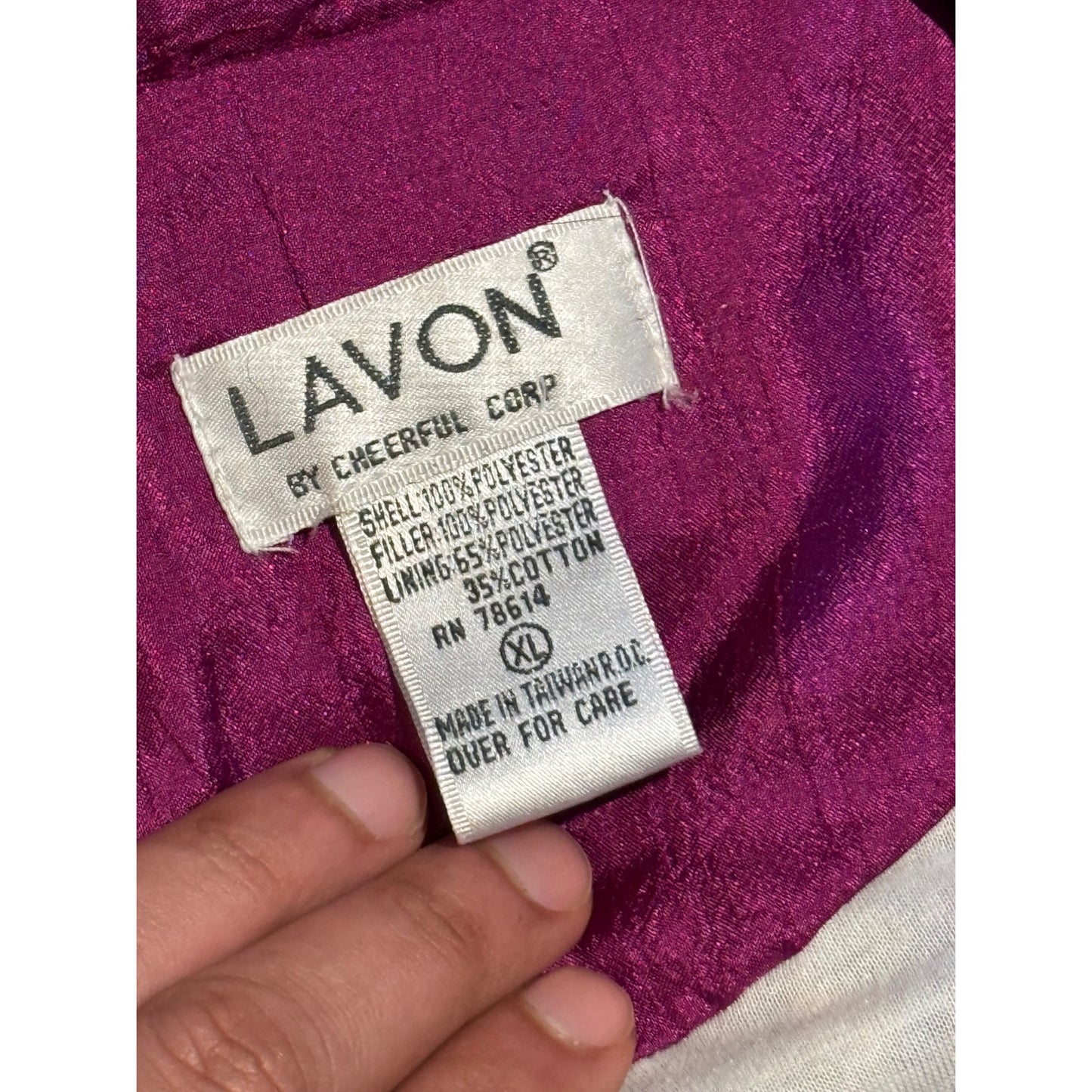 Close-up of a Lavon garment label sewn onto a magenta-purple fabric, with a hand holding the label in place. The visible text includes 'LAVON BY CHEERFUL CORP' and material details.