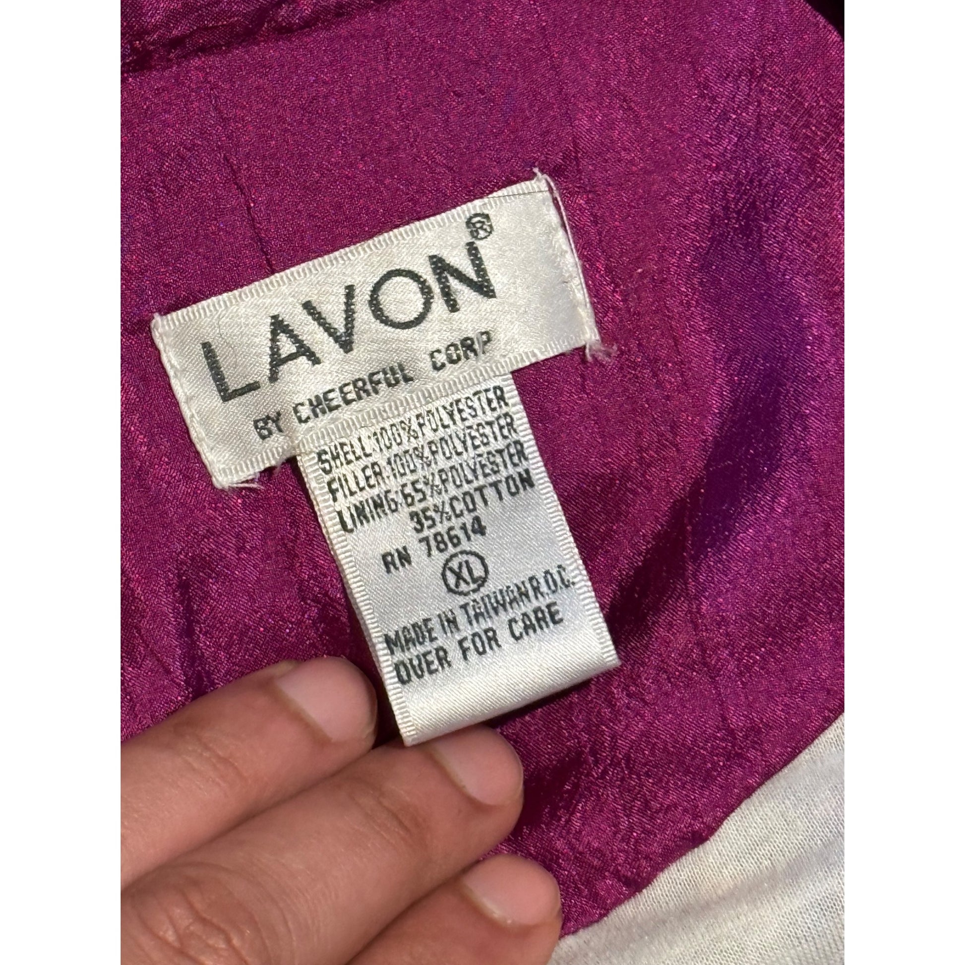 Close-up of a Lavon garment label sewn onto a magenta-purple fabric, with a hand holding the label in place. The visible text includes 'LAVON BY CHEERFUL CORP' and material details.