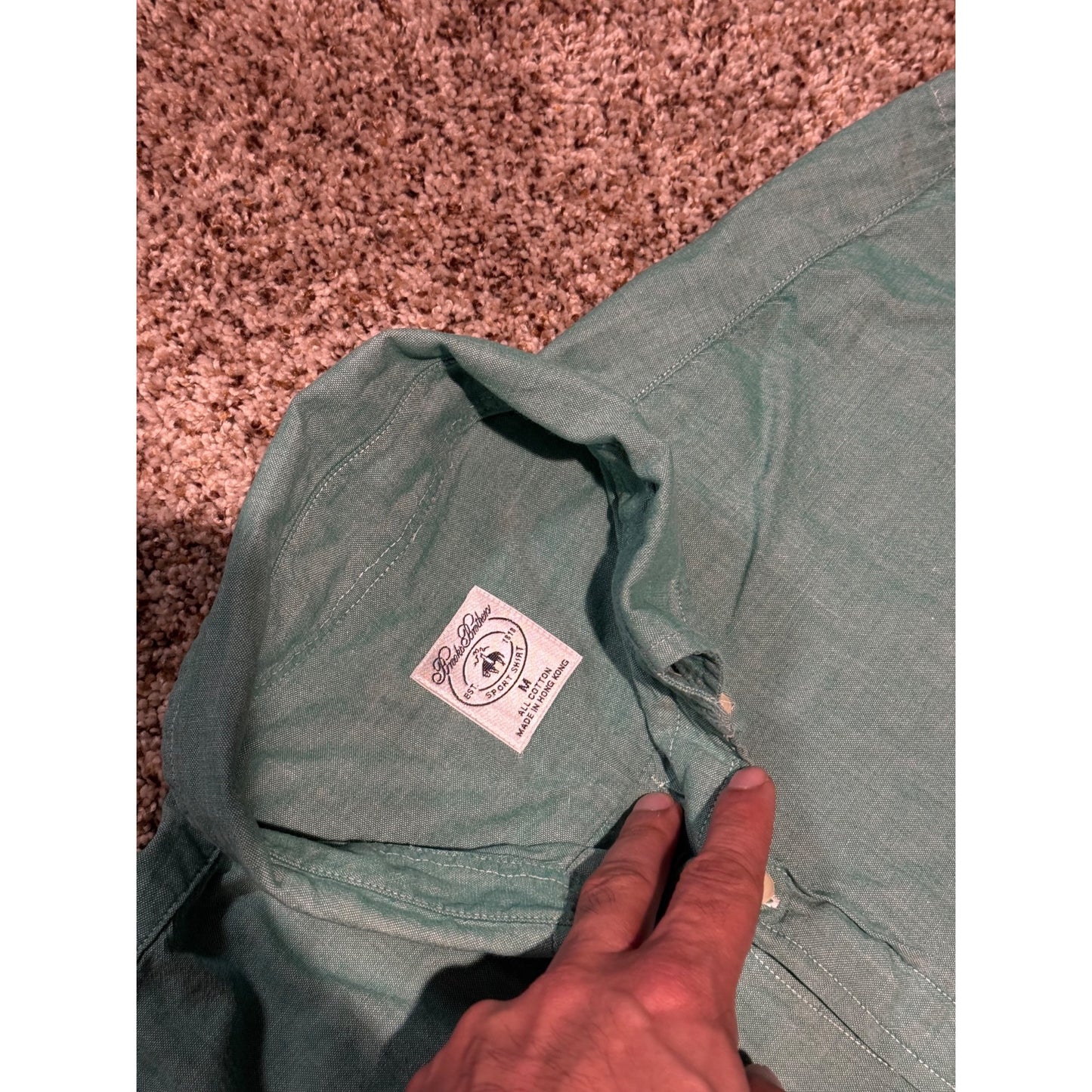 A close-up of a sage green denim shirt laid flat on a brown carpet, showing the inside collar with a white brand label and a hand pointing at it.