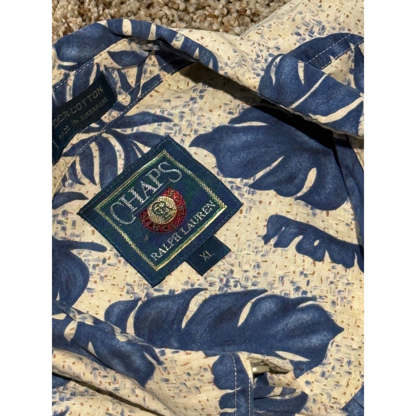 A close-up of a Ralph Lauren Chaps short-sleeve button-up shirt featuring a blue tropical leaf pattern on a cream speckled base. The Ralph Lauren patch logo is visible on the chest.
