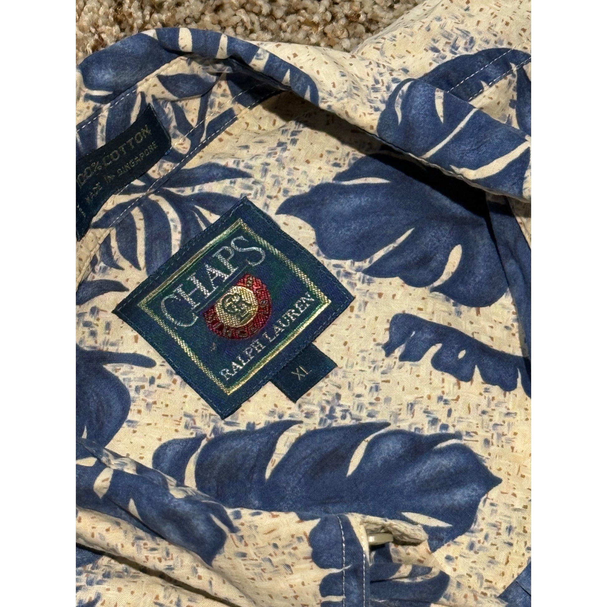 A close-up of a Ralph Lauren Chaps short-sleeve button-up shirt featuring a blue tropical leaf pattern on a cream speckled base. The Ralph Lauren patch logo is visible on the chest.