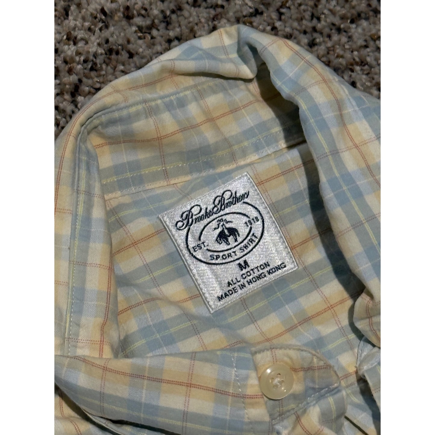 A close-up of a plaid button-up shirt collar with a visible 'Pierce & Barry' brand patch. The shirt features a light blue, beige, and yellow plaid pattern, photographed on a carpeted floor.