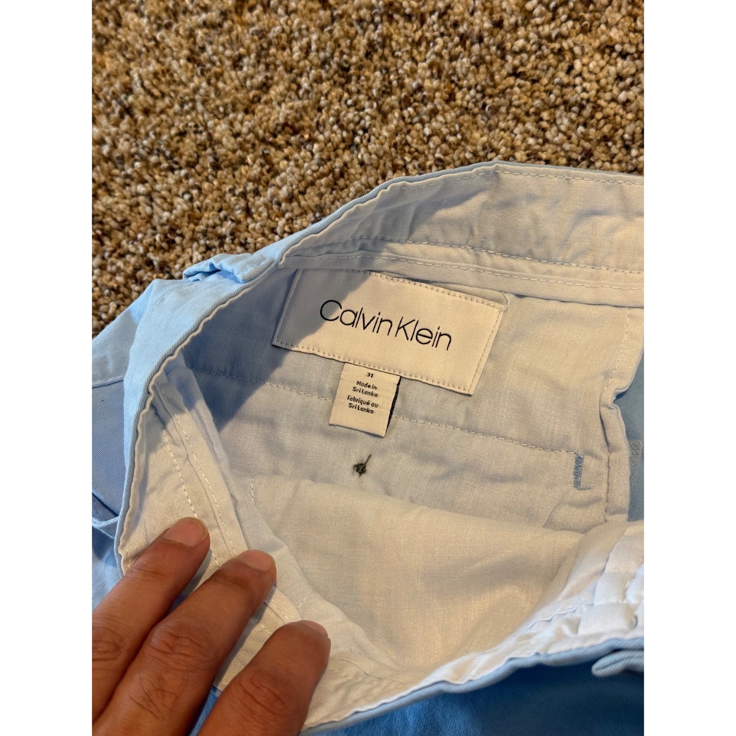 A close-up of light blue Calvin Klein pants showing the interior waistband and brand label, with a hand holding the fabric over a carpeted floor background.