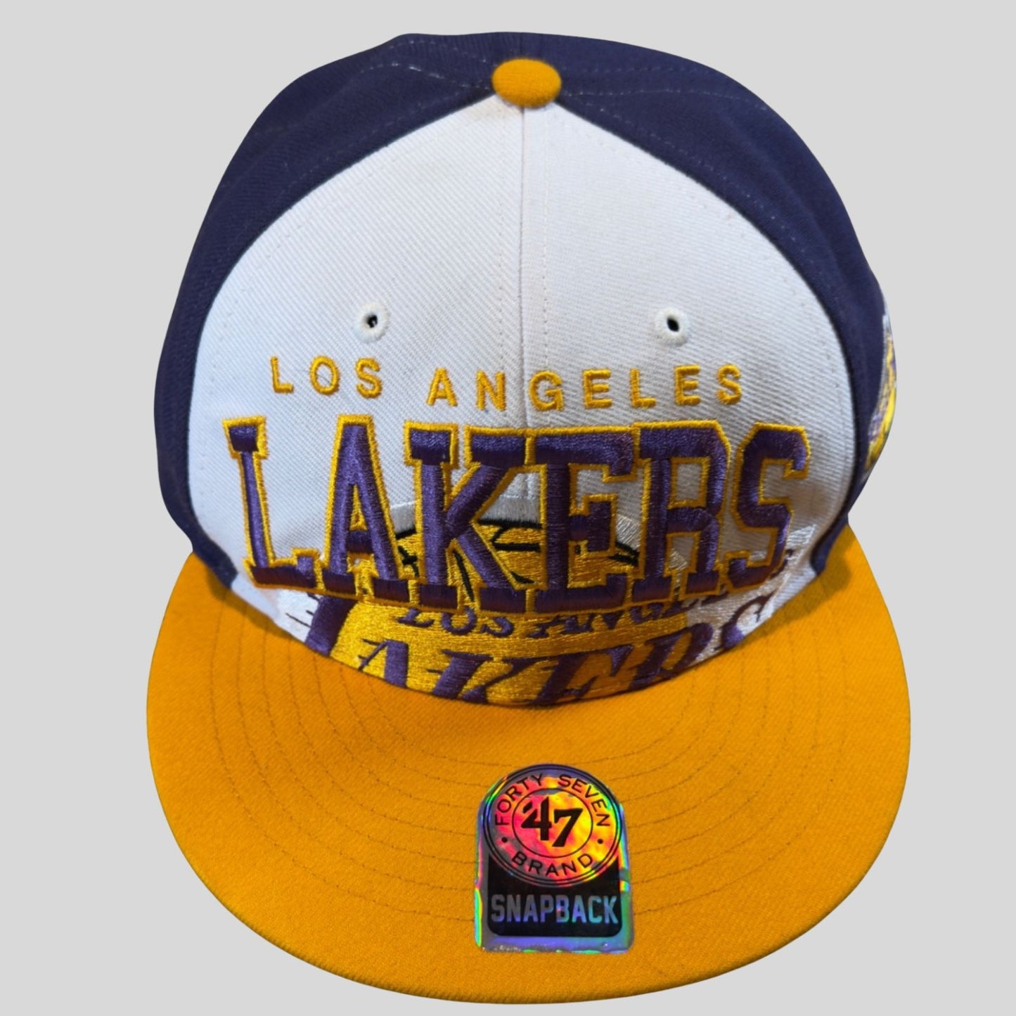 Los Angeles Lakers snapback cap with white front panel, yellow bill, and blue brim on a light gray background.