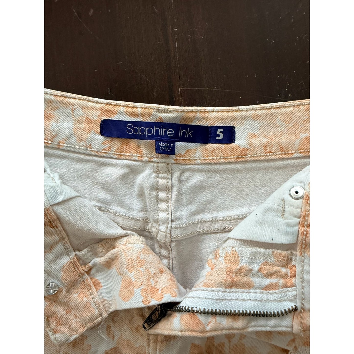 Close-up of the waistband of a pair of light beige denim jeans with a peach floral pattern, featuring a blue 'sapphire Ink' brand label.