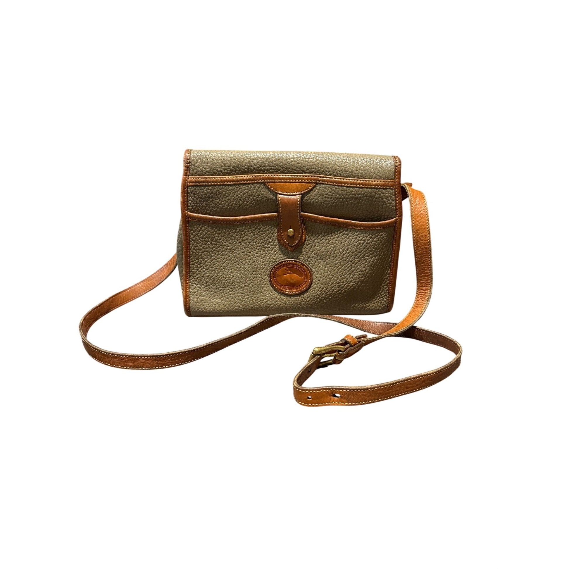 A small pebbled leather crossbody bag in olive green with tan leather trim and strap, featuring a front pocket and a circular metal logo medallion, shown on a plain white background.