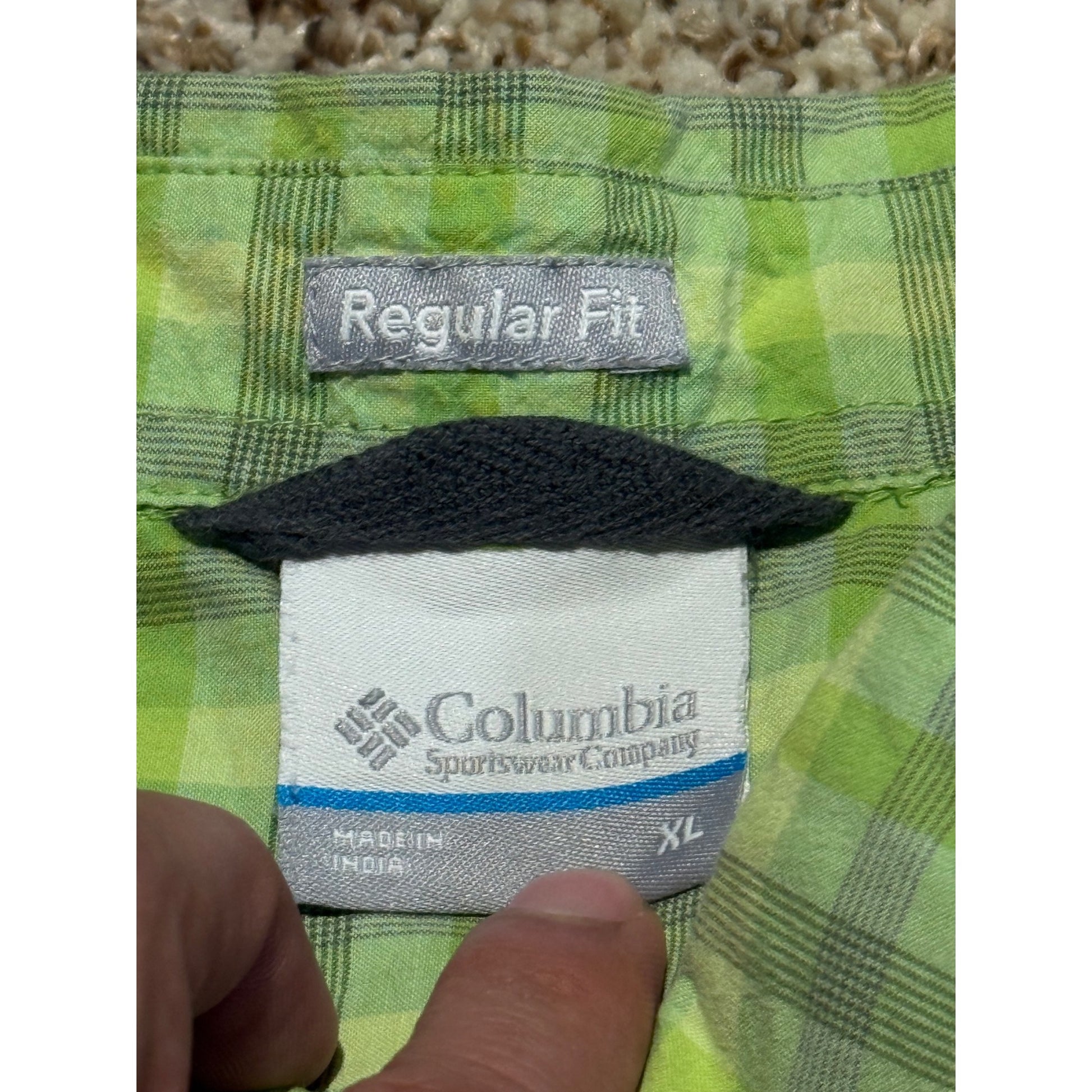 Close-up of a Columbia Sportswear Company garment label inside a green and yellow plaid garment, with a finger pointing at the label. The label reads 'Regular Fit' and 'XL'.