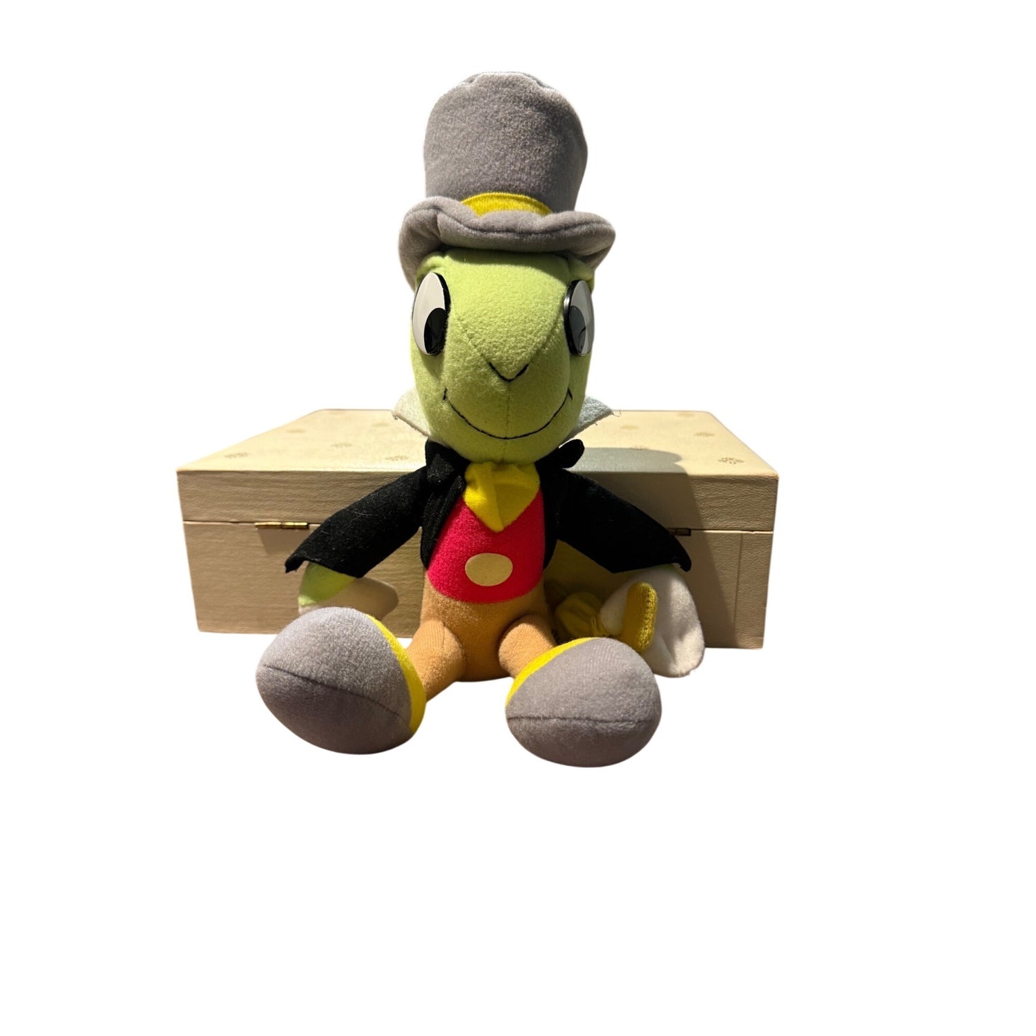 A plush toy of Jiminy Cricket wearing his signature top hat, black jacket, and red vest, sitting in front of a light wood crate on a white background.